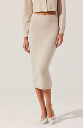 A woman is wearing a beige, knitted, long-sleeve cardigan with buttons, a matching beige, knitted midi skirt with a wide waistband, and white high-heeled shoes with a bow detail.